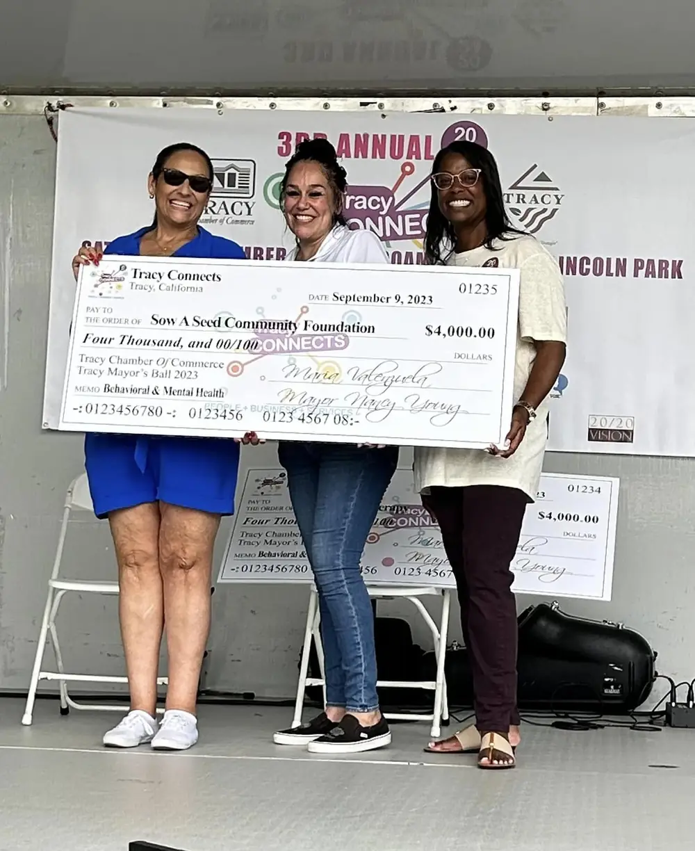 Three women on stage hold a $4,000 Tracy Connects check made out to Sow a Seed Community Foundation